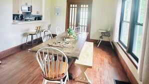 Ein Essbereich mit Holztisch, weißen Stühlen und einer modernen Pendelleuchte. Im Hintergrund ist eine Küche mit Schränken und Geräten zu sehen.