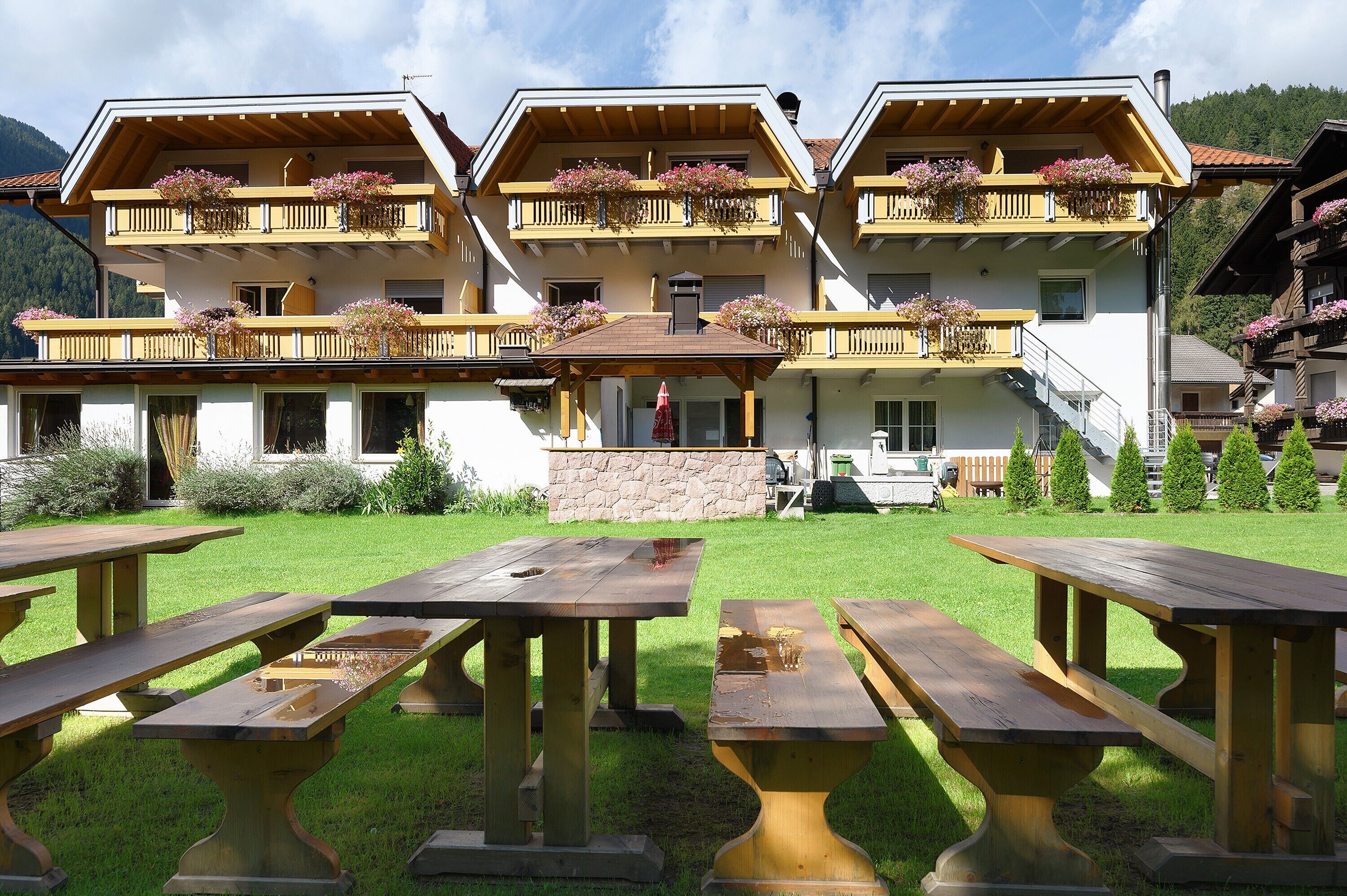 A hotel with multiple balconies adorned with flowers, a stone facade, and outdoor wooden picnic tables.