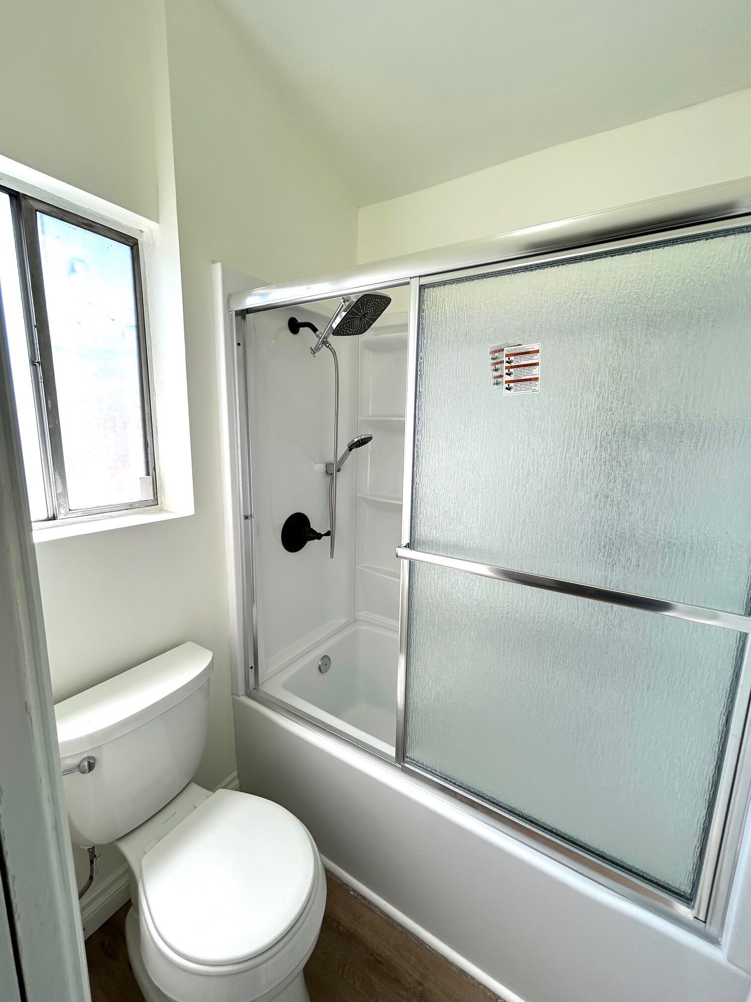 Un baño con inodoro, ducha con puerta de cristal y ventana sobre el inodoro.