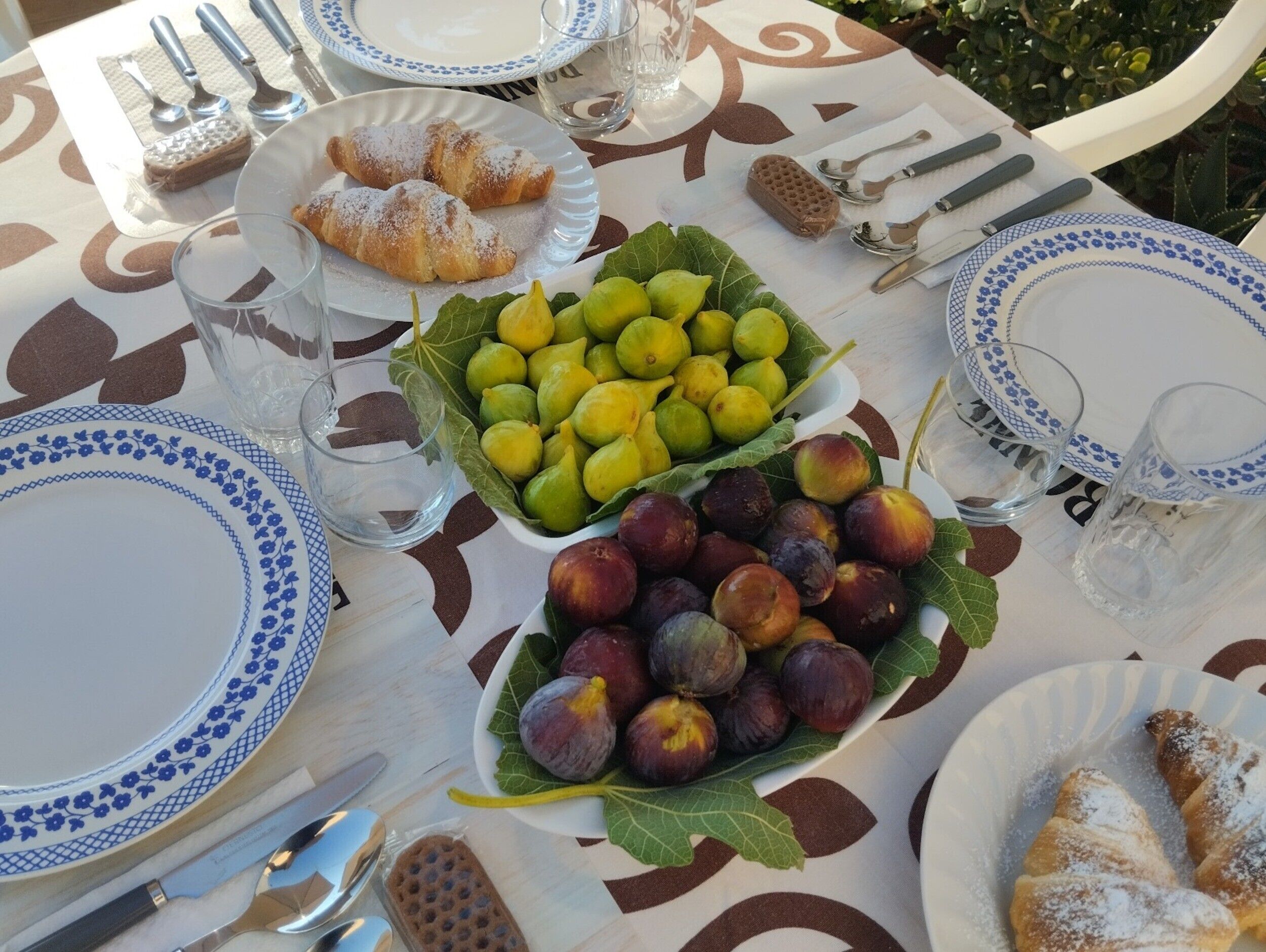 Una mesa dispuesta con fuentes de pasteles, higos y ciruelas, cubiertos y vasos.
