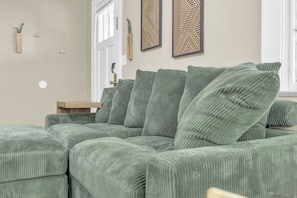 A green upholstered sectional sofa with a wooden side table, decorative wall art, and a window with blinds. - Spacious Stay w 2 King Beds Minutes to Downtown (Greenville)