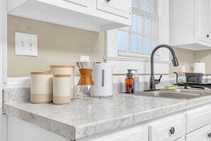 A kitchen with white cabinets, a marble countertop, a kettle, a coffee maker, a sink, and a window with blinds.
