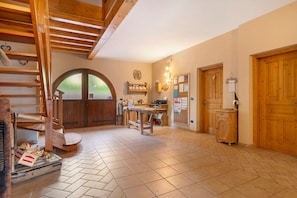 A rustic interior with a wooden staircase, a dining area with a table and chairs, a large arched doorway, and wooden doors.
