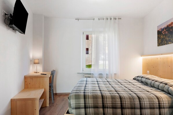 Ein Schlafzimmer mit Bett, Schreibtisch, Stuhl, Fernseher und einem Fenster mit Vorhängen.