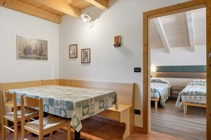 A room with a wooden table and chairs, a bed with a headboard, and a small wall-mounted shelf with flowers.