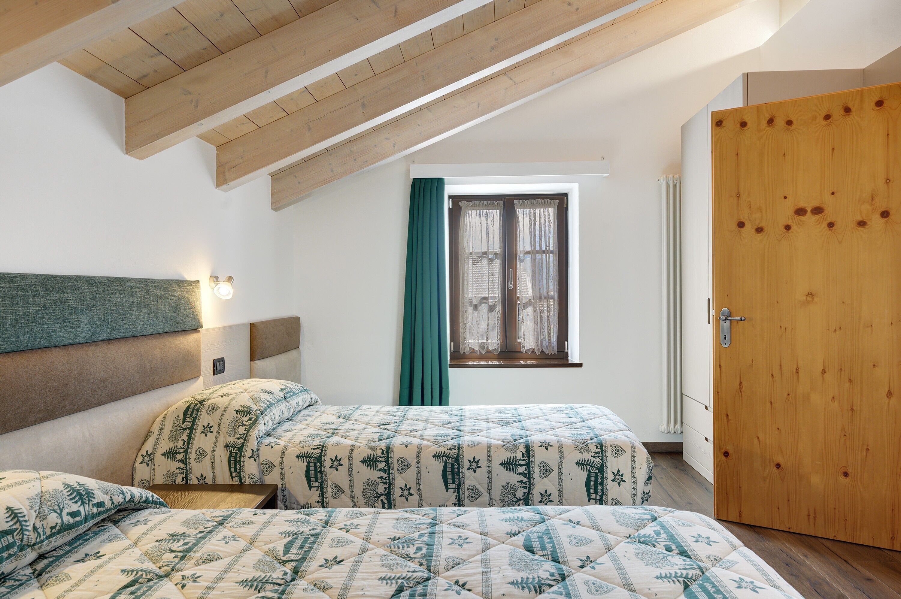 A bedroom with two beds, a window with curtains, a wooden door, and a sloped ceiling.