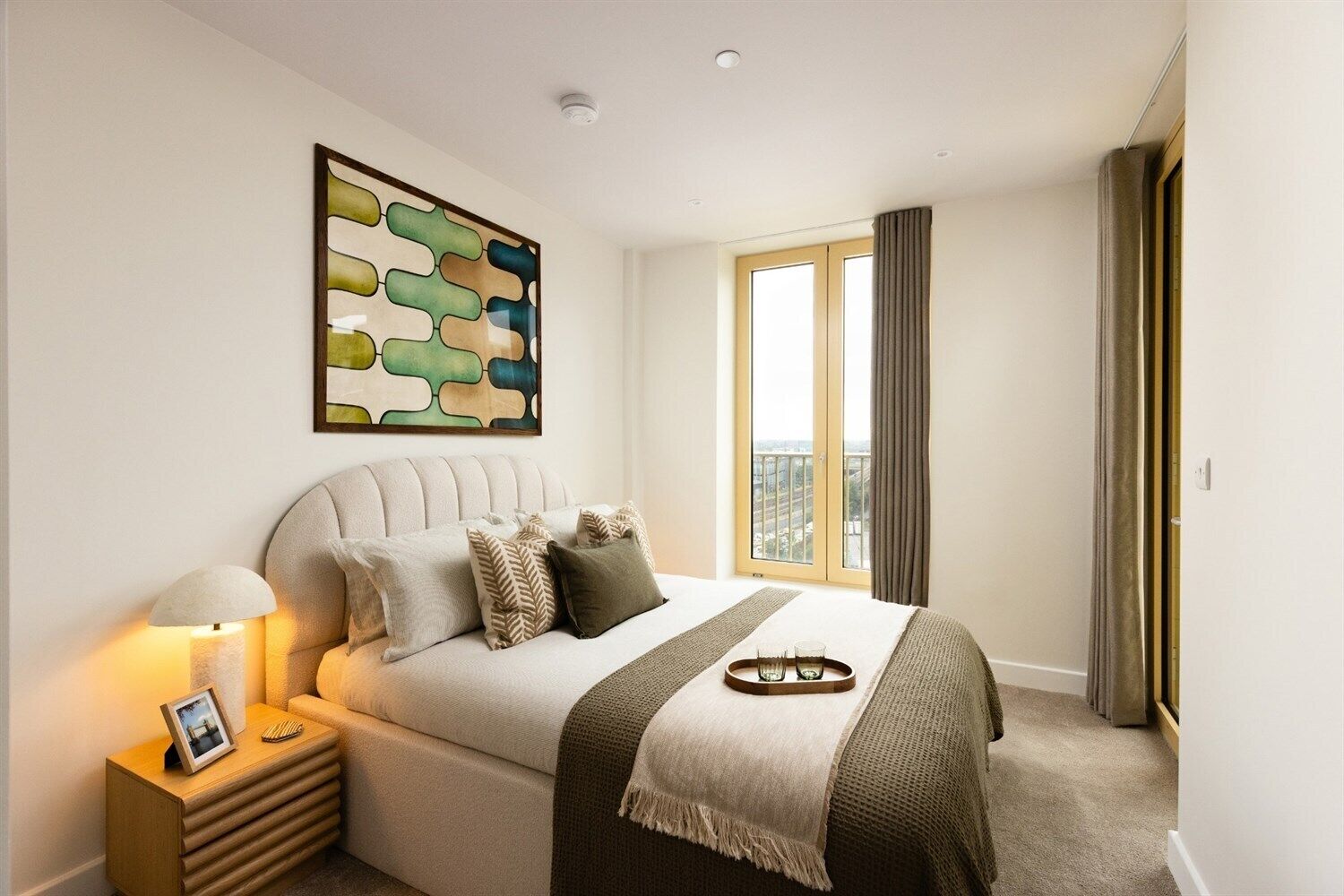 Quarto moderno com uma cama grande, mesa de cabeceira com abajur, quadro acima da cama e janela com cortinas.