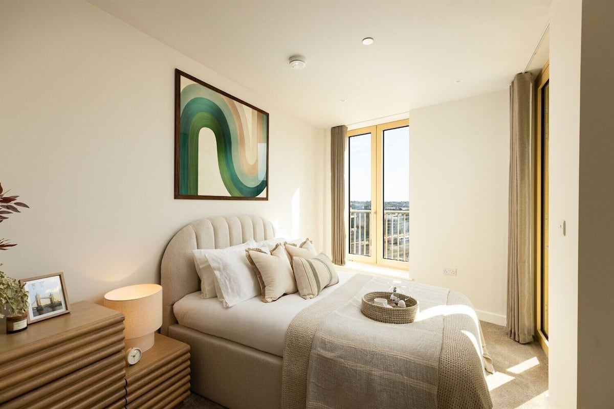 A modern bedroom with a large bed, a bedside table with a lamp, a framed picture, and a view of the city through a large window.