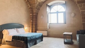 A room with a vaulted stone ceiling, a bed with a blue headboard, a small table, and a window with natural light. - King Suite with Private Jacuzzi - Charming in Felline | Borgo Don Ciccio (Felline)