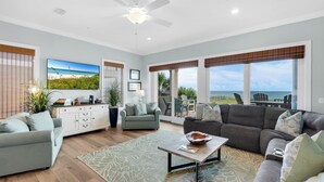 A spacious living room with a large window offering a view of the beach, a ceiling fan, and a variety of seating options including a gray sofa and a gray armchair. - Navarre Beachfront Townhome Southern Cross Sleeps 13 + Pet Friendly (Navarre)