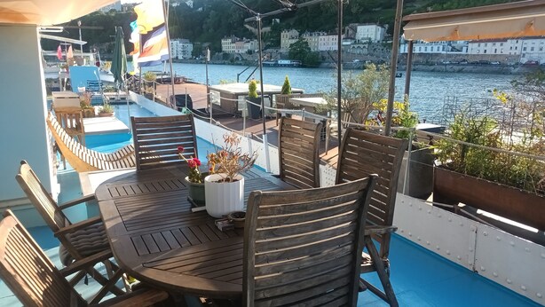 A deck with a round table, chairs, potted plants, and a view of the water and buildings. - ATYPICAL COMFORT ROOM ON A BARGE ON THE RIVER SAONE (Lyon)