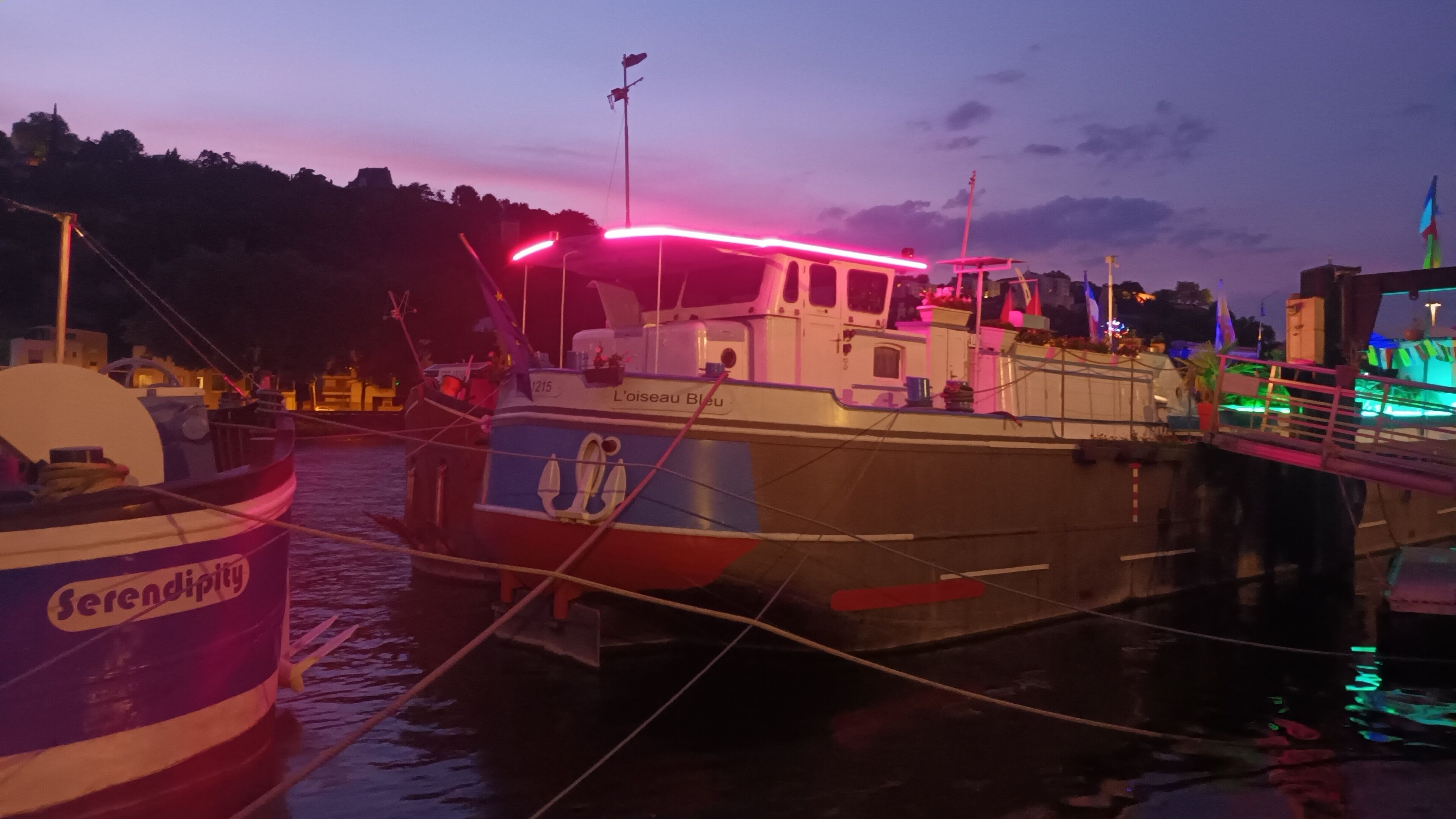 Ein Boot liegt in einer Marina festgemacht, mit dem Namen 'L'oiseau Bleu', und ein weiteres Boot trägt den Namen 'Serenopolis'.