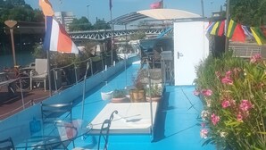 A pool area with a blue floor, lounge chairs, and a canopy. Flags are visible in the background. - ATYPICAL COMFORT ROOM ON A BARGE ON THE RIVER SAONE (Lyon)