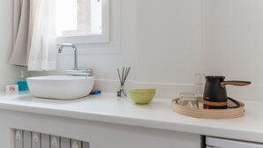 Une salle de bain avec un lavabo blanc, une fenêtre avec un rideau, un distributeur de serviettes, un distributeur de savon, un bocal en verre, un bol vert et une bouilloire posée sur un plateau.