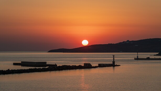 Un coucher de soleil sur une étendue d’eau calme, avec une colline lointaine et un phare.