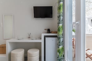 A modern room with a flat-screen TV, a small refrigerator, a white desk, two cushioned stools, and a door leading to an outdoor area.