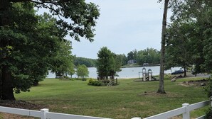 Un'area verdeggiante con una recinzione bianca, alberi e un lago sullo sfondo, dove si intravede una casa e un'area giochi.