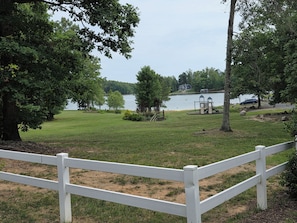 A grassy area with a white fence, trees, and a lake with a house and playground in the background.