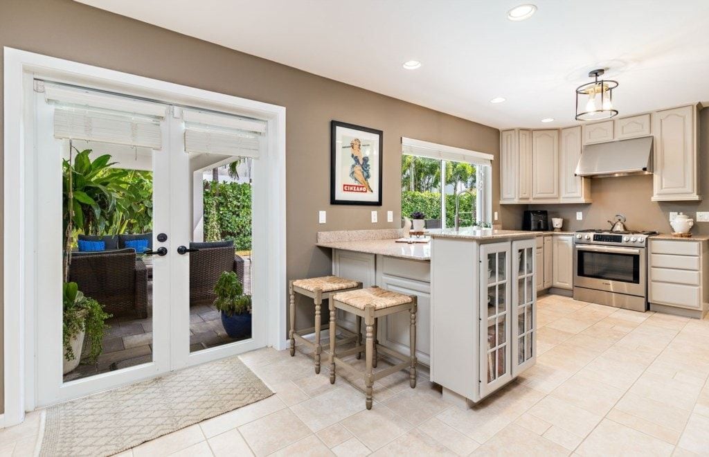 A kitchen with a central island, a stove, and a sink. There are two stools and a door leading to an outdoor area with plants.
