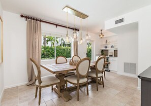 A dining area with a wooden table and chairs, a hanging light fixture, and a view of greenery outside.
