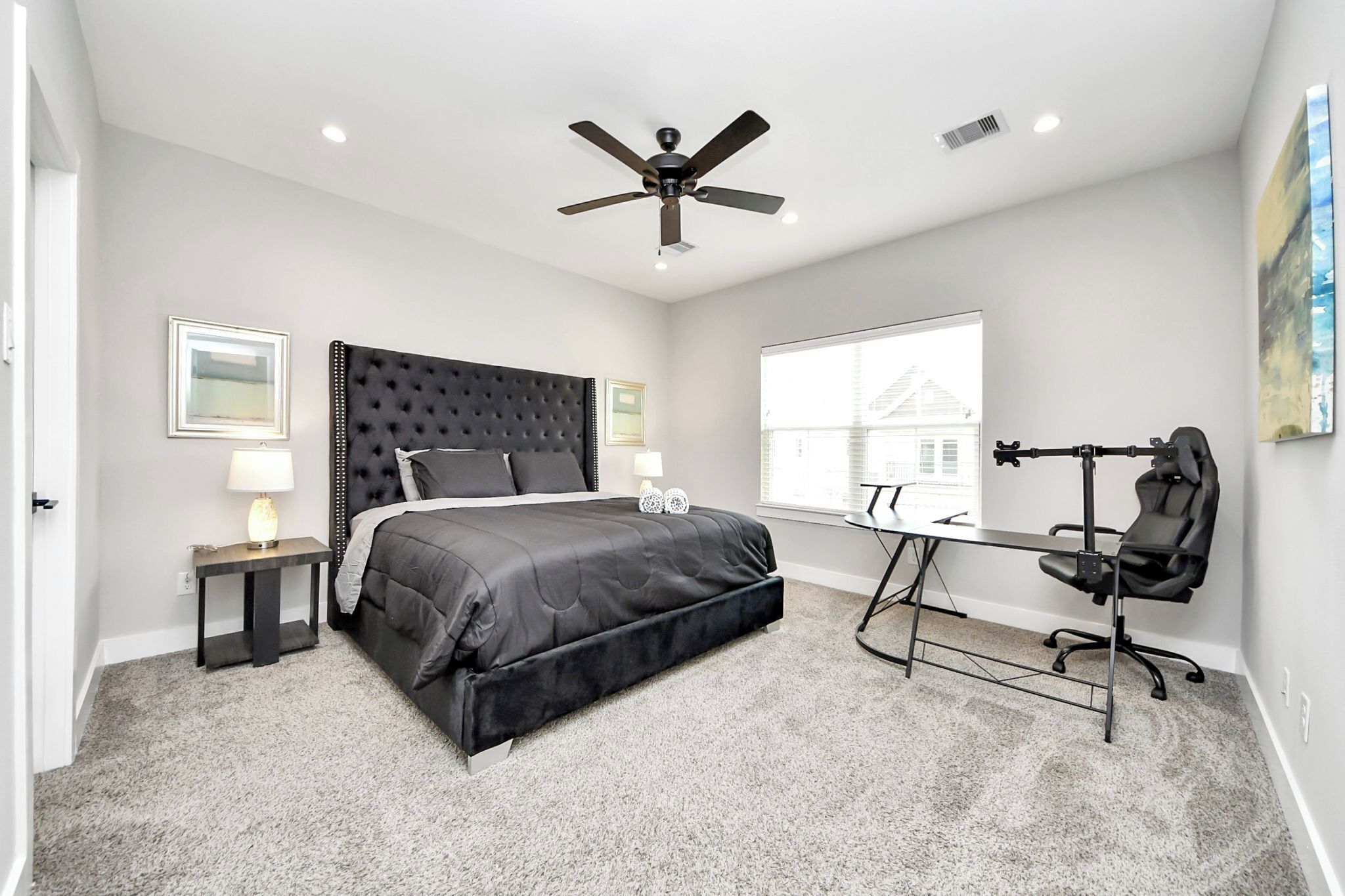 A bedroom with a large bed, a desk, a chair, a ceiling fan, and a window with a view of a house.