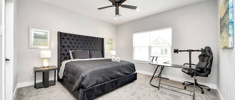 A bedroom with a large bed, a desk, a chair, a ceiling fan, and a window with a view of a house.