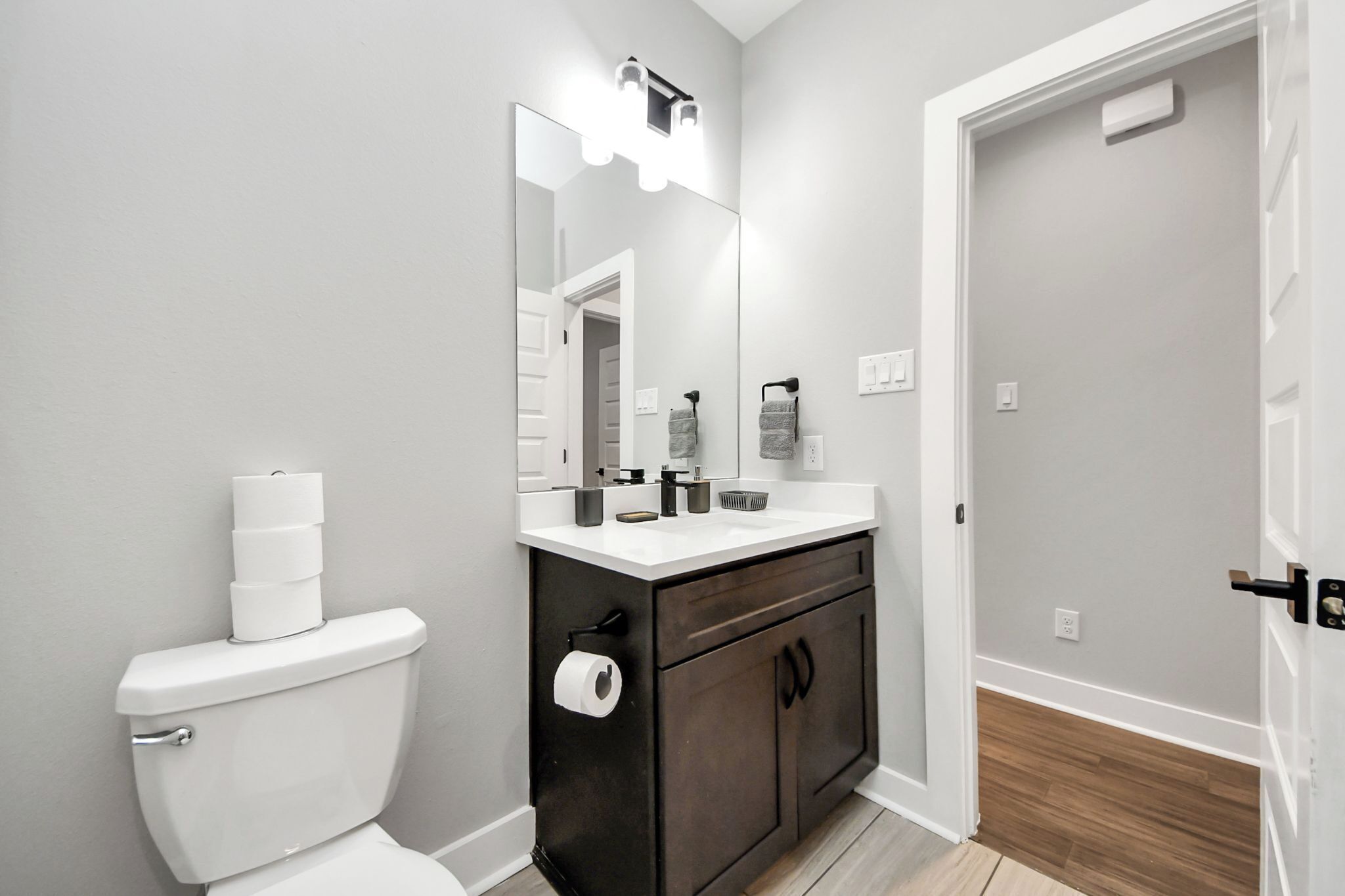 Ein modernes Badezimmer mit Toilette, dunklem Holzschrank, weißer Arbeitsplatte und einem beleuchteten Spiegel.