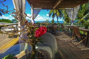Un área de comedor al aire libre con vista al océano, muebles de madera y una mesa de vidrio con un arreglo floral.