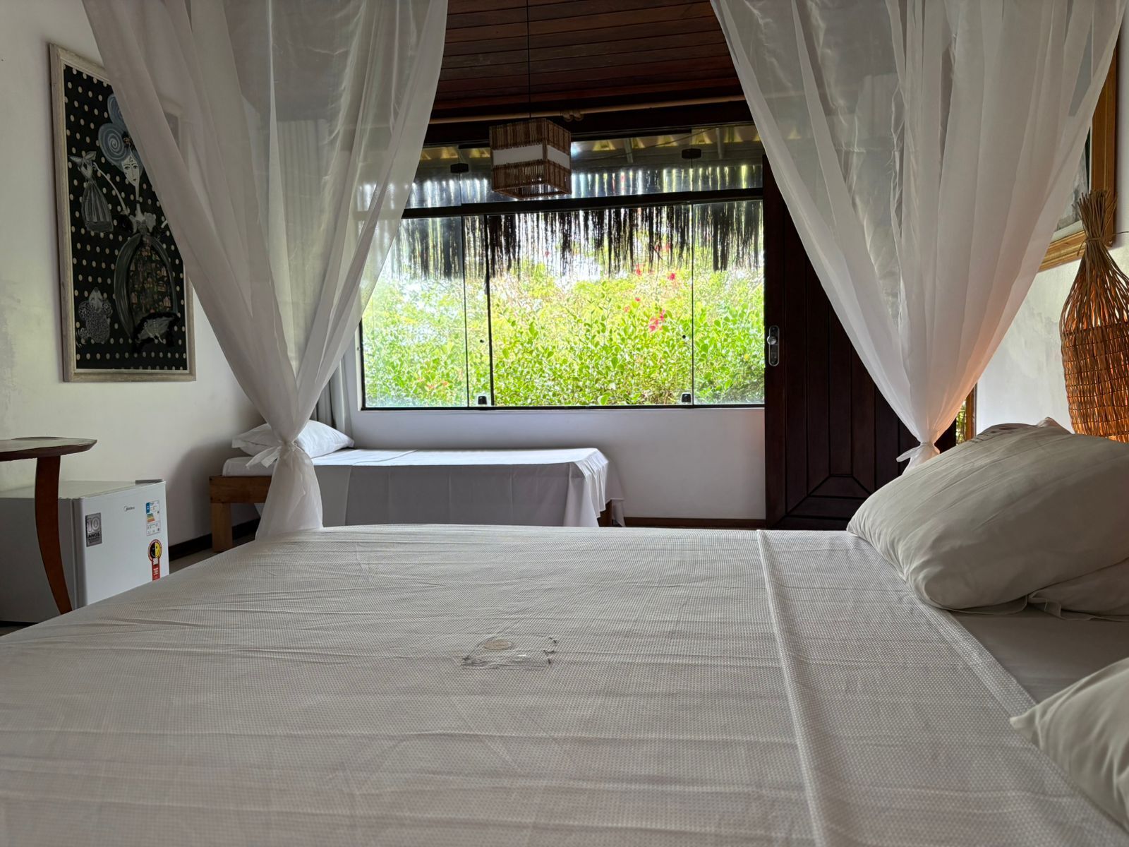 A bedroom with a bed, a window with curtains, a small table, and a wall-mounted picture.