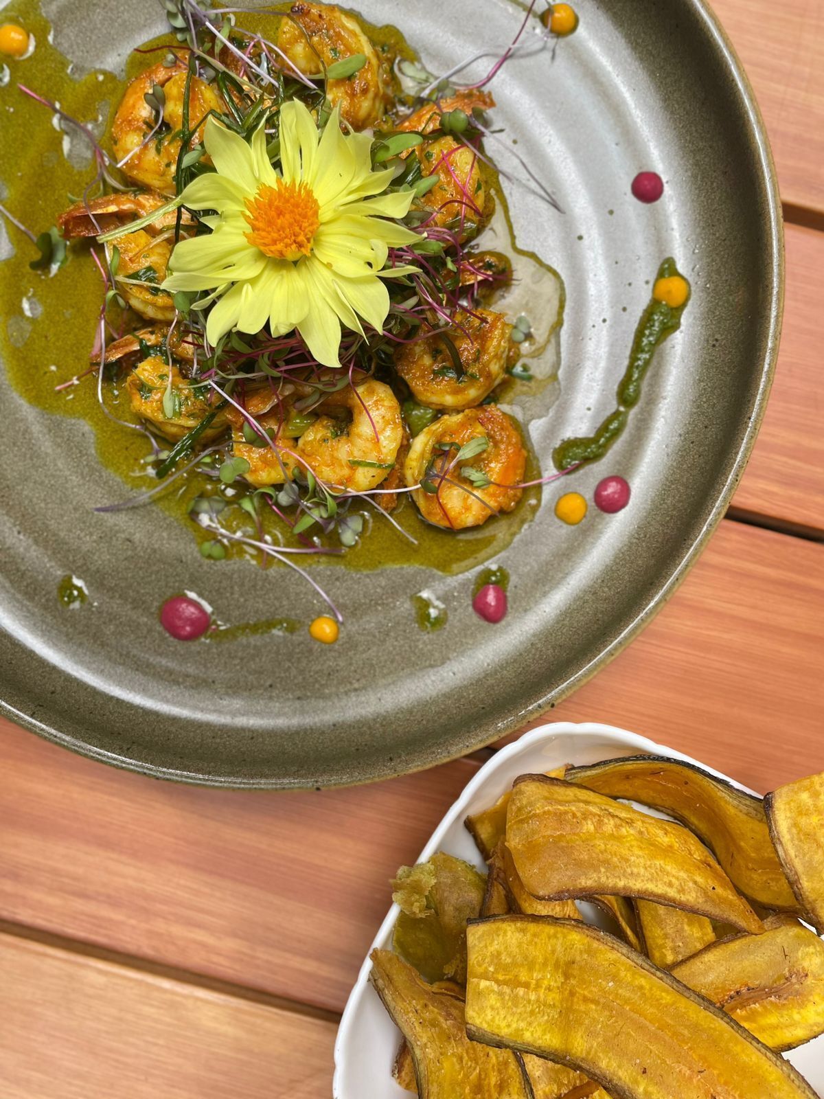 Une assiette de nourriture comprenant des crevettes et une fleur décorative, ainsi qu’un bol de bananes plantains frites.