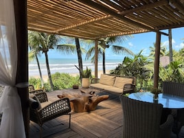 Una terraza de madera con mobiliario de mimbre, una mesa de vidrio y vista a la playa y al océano.