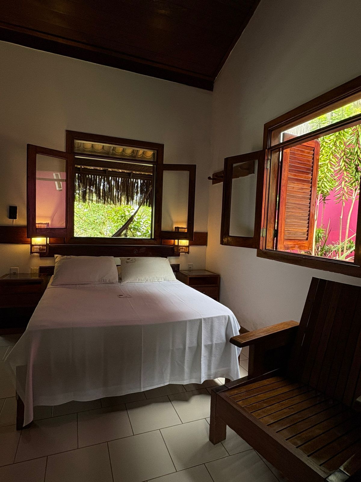 A bedroom with a bed, two nightstands, a wooden chair, and a window with a view of greenery.