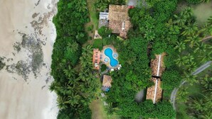 An aerial view of a resort with a swimming pool surrounded by lush greenery and palm trees, a beach, and several thatched-roof structures. - Pousada Morro dos Navegantes (Ilhéus)