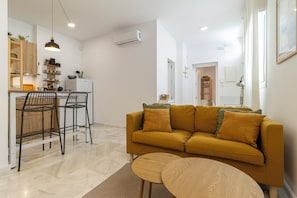 Ein modernes Wohnzimmer mit Sofa, Essbereich mit Stühlen, Küche inklusive Kühlschrank und Klimaanlage.