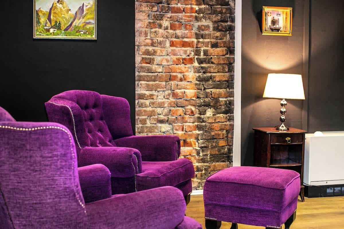 Quarto com poltronas estofadas em roxo e um banquinho do mesmo tom, parede de tijolos, quadro emoldurado e abajur.