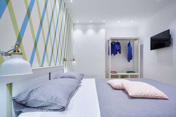 A modern bedroom with a bed, a bedside lamp, a wardrobe with hanging clothes, and a TV mounted on the wall.