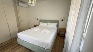 A neatly made bed with white linens and towels, a wooden nightstand with a lamp, and a door with an air conditioning unit.
