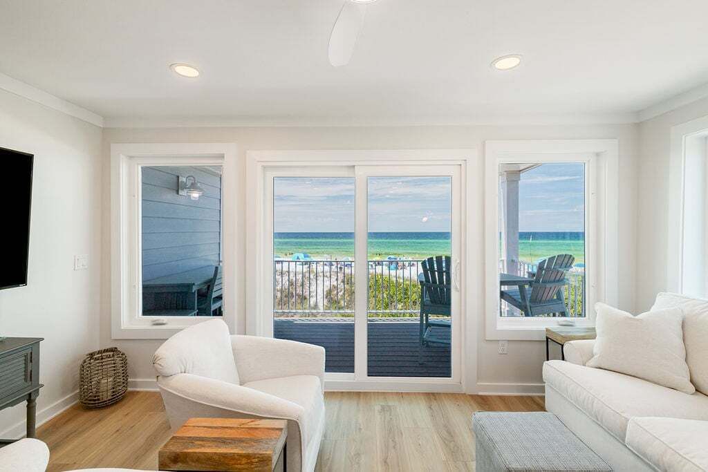 A room with a large sliding glass door leading to a balcony with a view of the ocean. There are two white chairs and a wooden table on the balcony.