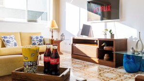Ein Wohnzimmer mit einer Couch, einem Couchtisch mit Popcorn und Coca-Cola-Flaschen, einem Fernseher, der 'NETFLIX' zeigt, und einer Holzkommode.