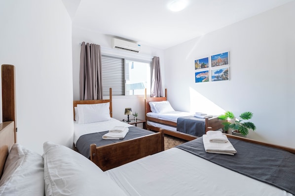 A hotel room with two single beds, a window with curtains, a wall-mounted air conditioner, and a small table with a lamp.