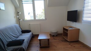 A small, sparsely furnished room with a wooden floor, a grey sofa, a small wooden table, a grey ottoman, a radiator, a TV mounted on the wall, and a window with blinds.