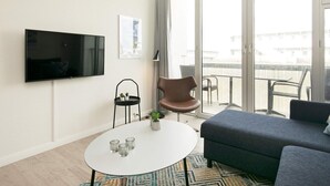 A modern living room with a flat-screen TV, a round coffee table with glasses, a brown chair, a grey sofa, and a balcony with outdoor furniture.