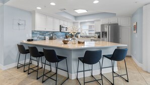 Una cocina moderna con una isla grande, taburetes de bar, armarios blancos y electrodomésticos de acero inoxidable.