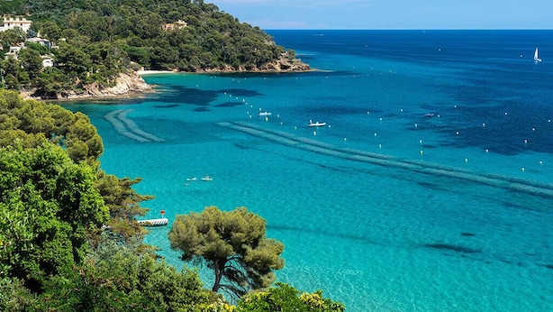 A coastal landscape with clear turquoise waters, boats, and a tree-lined hill.