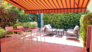 Een terras buiten met een gestreept zonnescherm, een tafel met stoelen en een haag op de achtergrond.