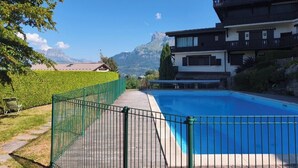 A swimming pool with a view of mountains, surrounded by a fenced area and a building with balconies.