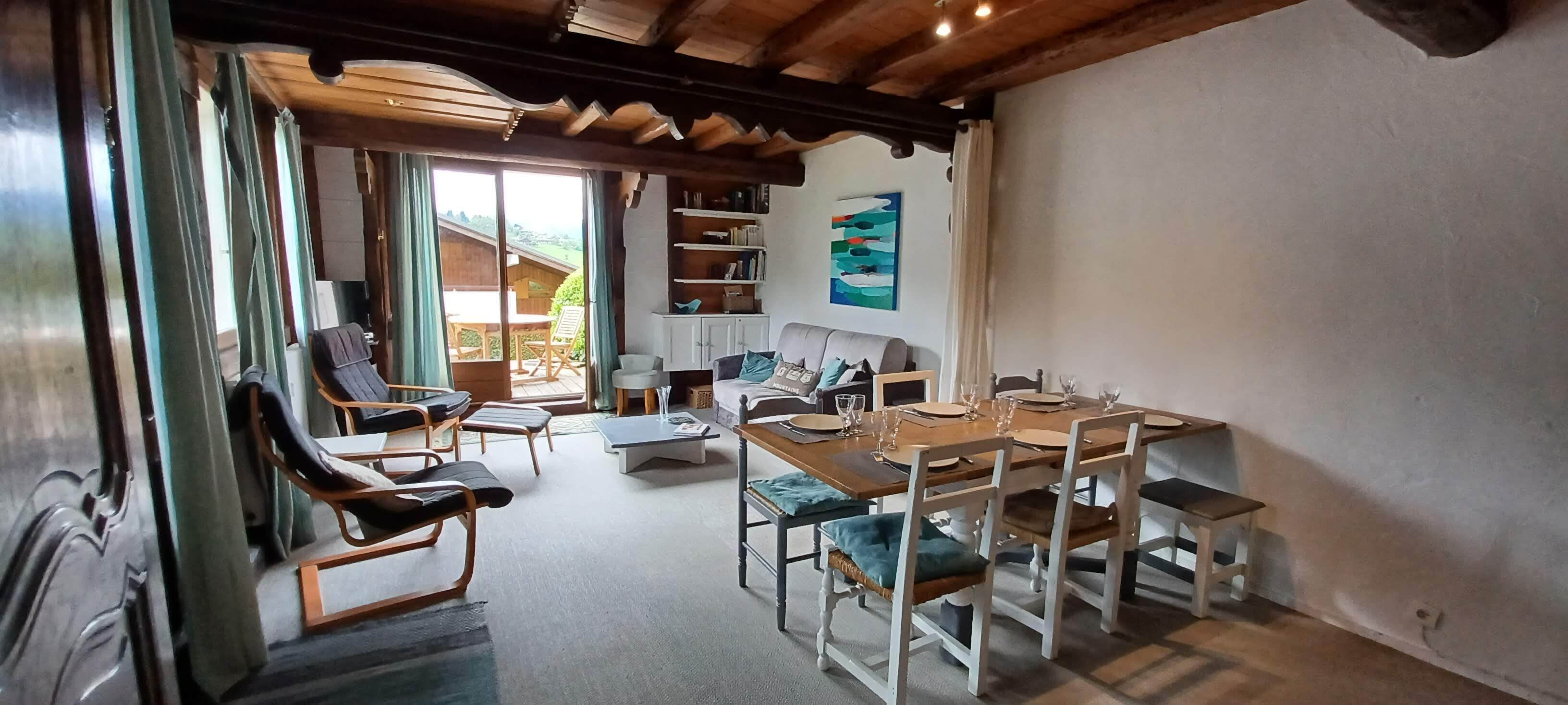 A cozy dining area with a wooden table set for four, surrounded by white chairs with cushioned seats. A sofa and a small table with a vase are in the background.