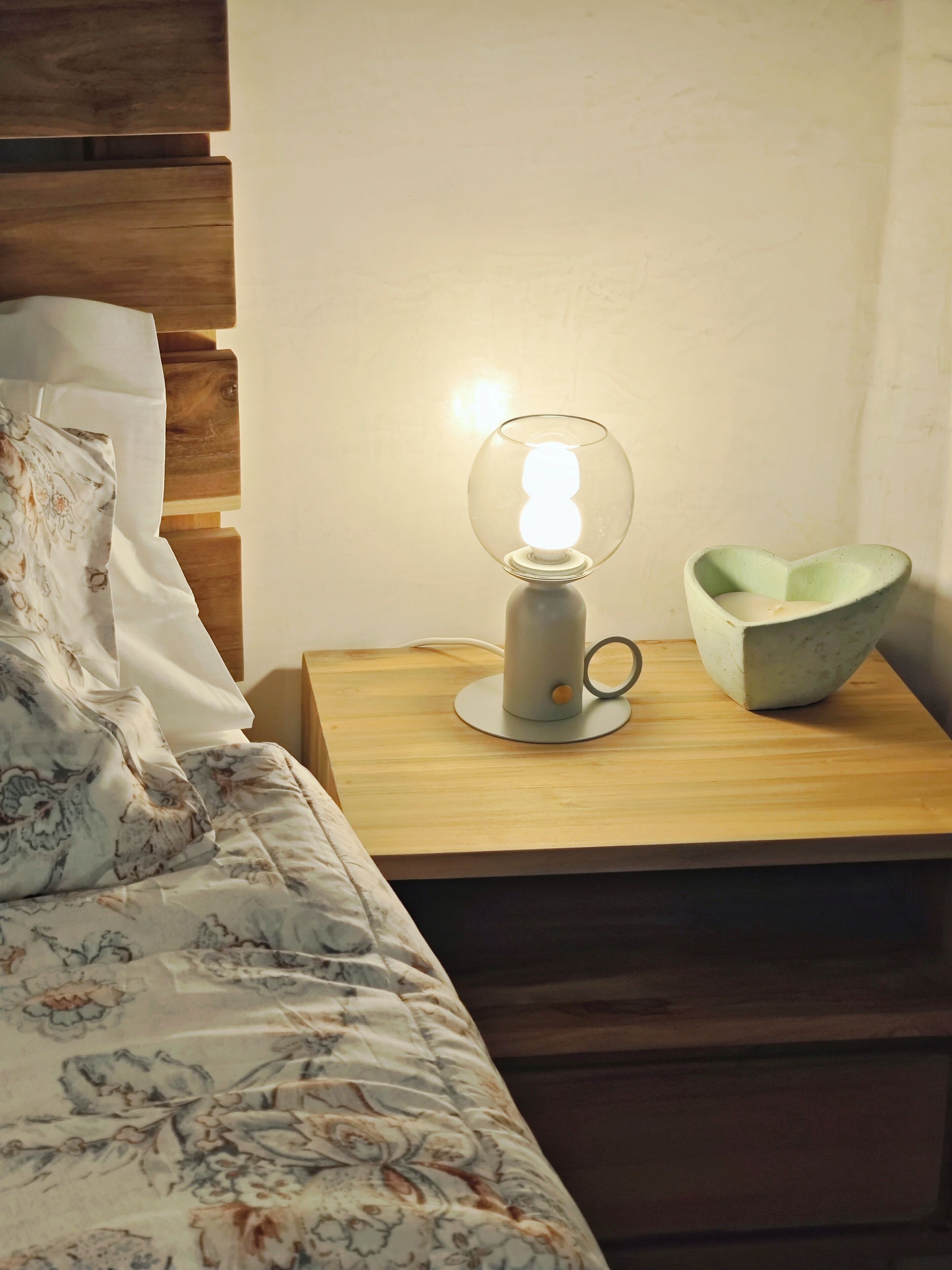 Un lit bien fait, avec une couette à motifs floraux, une table de chevet sur laquelle se trouvent une lampe et un bol en céramique, et une tête de lit en bois.