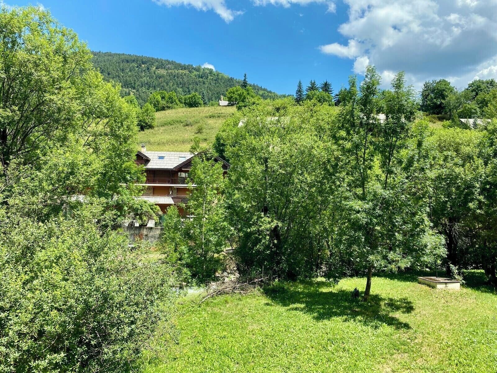 A wooden cabin with a balcony, surrounded by lush greenery and a hill in the background.