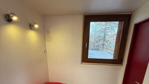 Une petite chambre avec un lit simple, un oreiller rouge, une fenêtre donnant sur les arbres et des lampes fixées au mur.